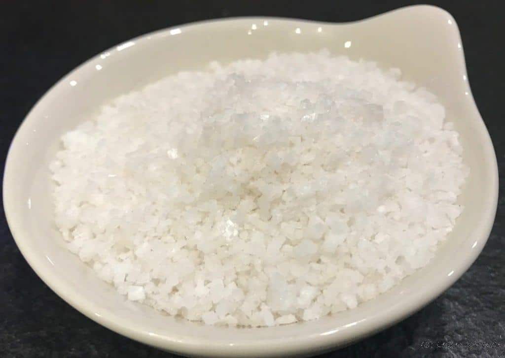 Grobe Salzkristalle mit einer Kantenlänge von ca. 2 mm in einer weoßen Porzellanschüssel vor schwarzem Hintergrund.