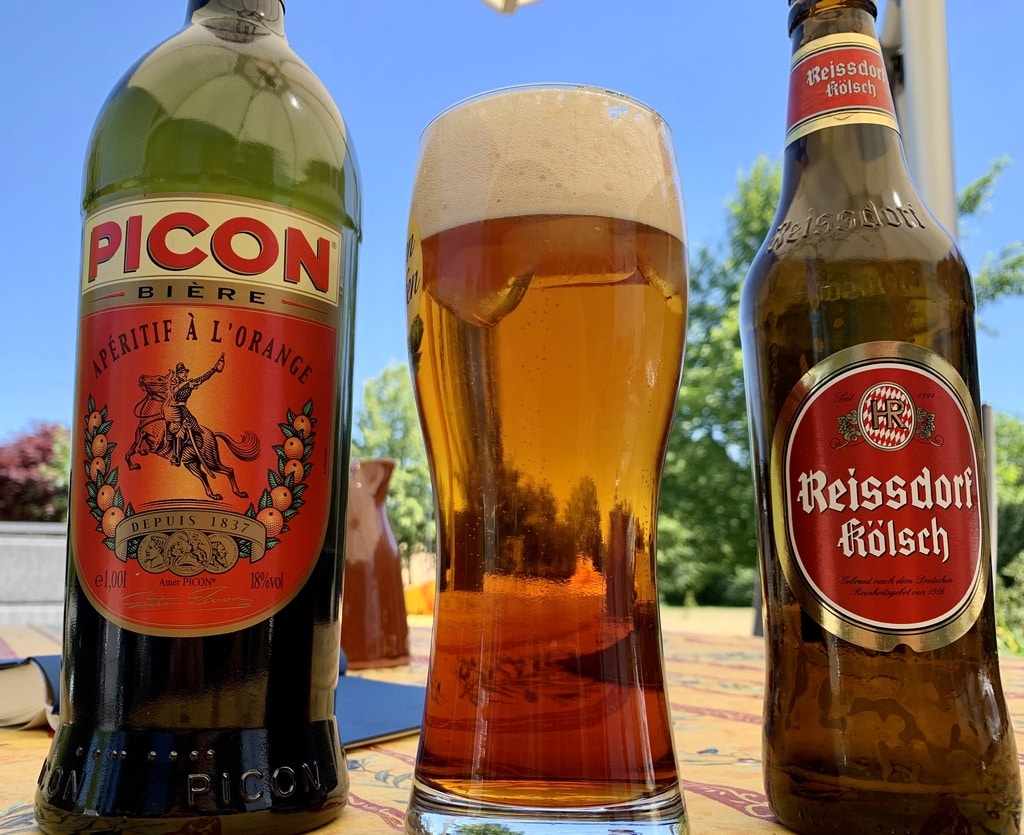 Eine Flasche Picon, ein Glas mit Picon Bière und daneben eine Flasche Kölsch vor blauem Himmel.