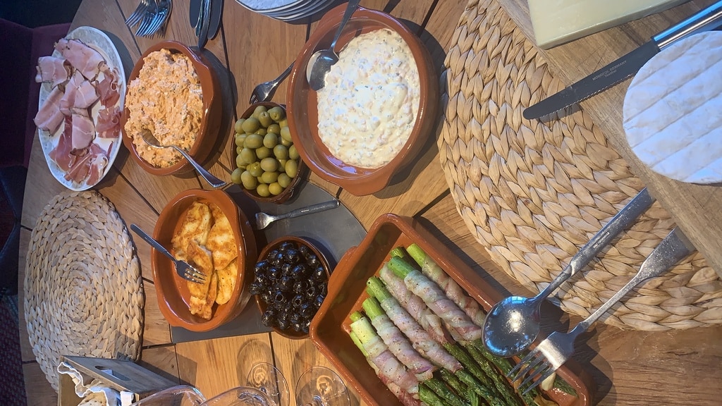 Ein Holztisch mit vielen Tapas wie z.B. grüne und schwarze Oliven, Jamon, Spargel im Baconmantel, Käse und ein paar Cazuelas. Schön zusammengestellt für einen bunten Tapas Abend.
