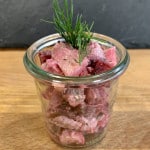 Ein kleines Weckglas gefüllt mit Matjessalat und dekoriert mit einem Stängel Dill auf einer Holzplatte vor schwarzem Hintergrund.