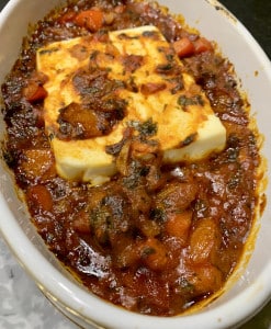 Rezept für Feta Saganaki - eine leckere griechische Vorspeise mit geschmortem Gemüse, Kräutern und Schafskäse. Auf dem Bild ein überbackener Schafskäse auf geschmorten Tomaten und Paprika in einer weißen Auflaufform, frisch aus dem Backofen.