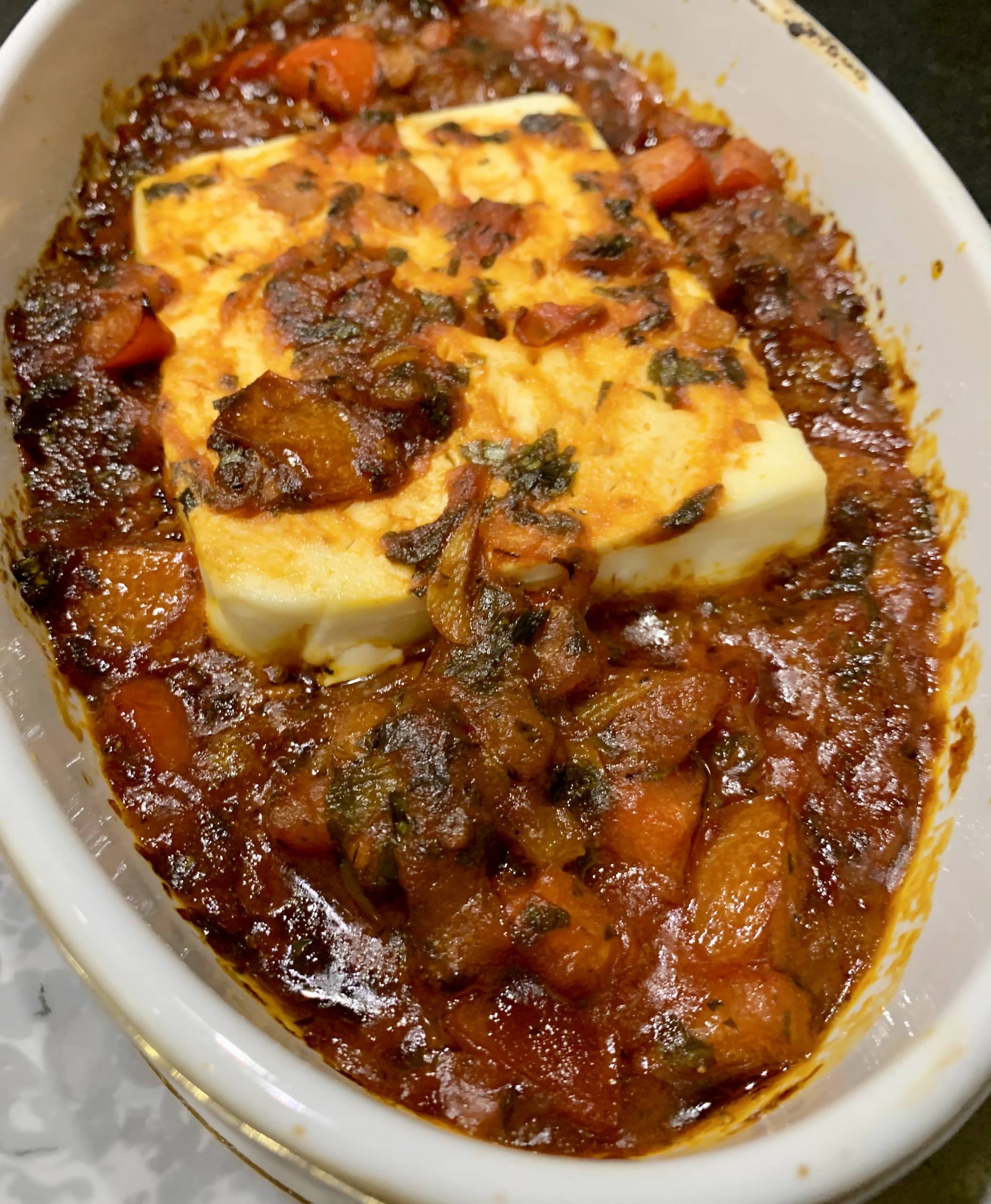Rezept für Feta Saganaki - eine leckere griechische Vorspeise mit geschmortem Gemüse, Kräutern und Schafskäse. Auf dem Bild ein überbackener Schafskäse auf geschmorten Tomaten und Paprika in einer weißen Auflaufform, frisch aus dem Backofen.