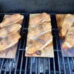 Drei Zedernholzbretter auf einem Grillrost, darauf befinden sich Lachsfilets, die gerade frisch als Flammlachs fertig geworden sind.