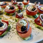 Auf einem großen weißen Porzellanteller sind Tomatenstückchen auf Gurkenscheiben mit Zwiebelringen, schwarzen Oliven und einem Stück Feta einzelnen angerichtet. Das ´Ganze ist mit einem Kräuter-Essig-Öl Dressing beträufelt und stellt einen neu interpretierten Griechischen Bauernsalat dar.