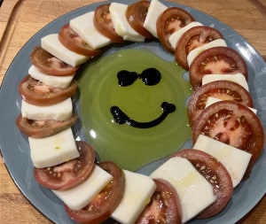 Tomate Mozzarella im Kreis auf einem blauen Teller angerichtet und mit Olivenöl-Balsamico Spiegel als Smile in der Mitte.