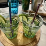 Zwei Gläser Margarita, ein Cocktail Klassiker, in grünen Gläser mit schwarzem Strohhalm, Salzrand und Limettenscheibe auf einem runden Holzbrett. Im Hintergrund Holzstößel, Eiswürfelzange, Cocktailshaker, ein leeres blaues Glas sowie Barzubehör leicht verschwommen.
