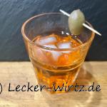 Ein Glas Aperol Spritz mit Eiswürfeln und Olive auf einem Zahnstocher auf einem Holzbrett vor einer Schiefertafel-