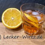 Ein Glas Aperol Spritz mit Eiswürfeln und Orangenscheibe auf einem Holzbrett vor einer Schiefertafel- Daneben eine halbe Orange.