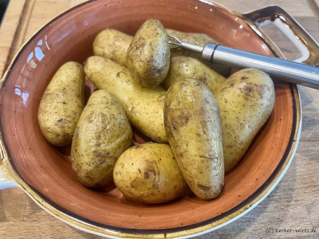 In einer pastell-roten Porzellanschale liegen elf Pellkartoffeln und eine dreizackige Kartoffelgabel.