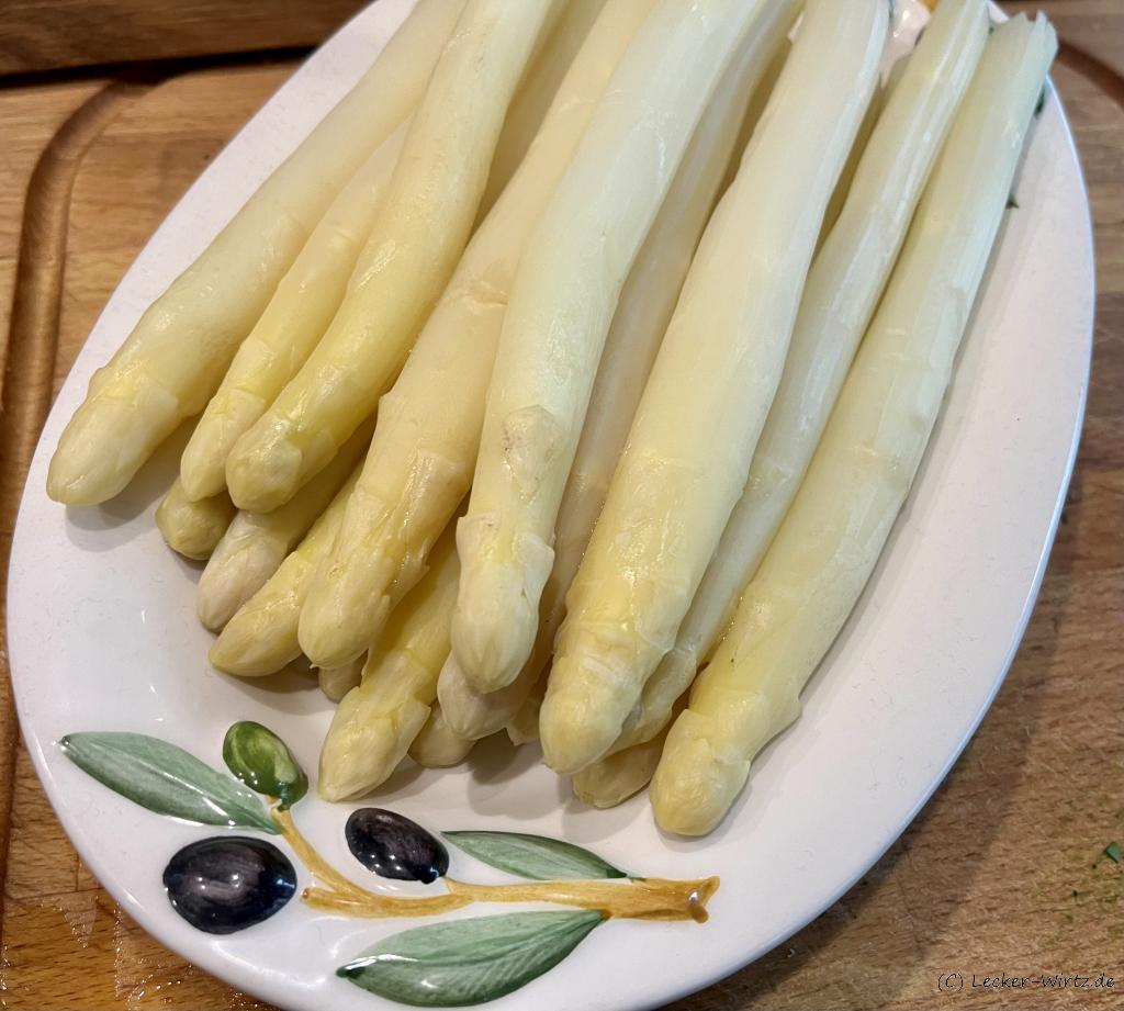 Eine ovale Porzellanschüssel mit mehreren Stangen gekochtem Spargel auf einem Holzbrett.