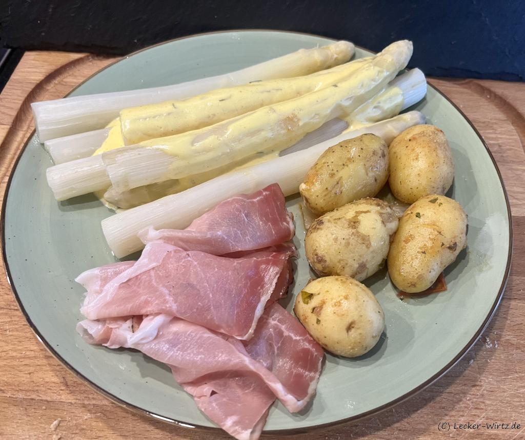 Sieben Stangen gekochter weißer Bornheimer Spargel auf einem pastell-grünen Teller mit 3 Scheiben rohem Schinken und fünf Pellkartoffeln mit Schale.