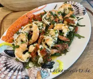 Auf einem bunten, ovalen Teller im Seafood Style sind Garnelen auf einem Bett aus Quelle. Das Gericht aus Frankreich heißt Crevettes grillées au beurre.