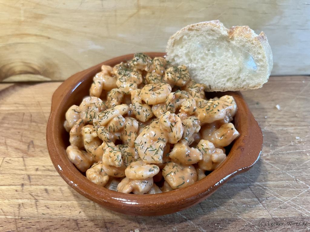 Ein Garnelencocktail in einer Cazuela mit Dill bestreut und einem Stück Baguette auf einer Holzplatte.