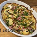Eine weiße Auflauf form mit Poêlée normande ( normannische Bratkartoffeln mit Camembertsauce) frisch überbacken aus dem Backofen auf einem Holzbrett. Garniert mit frisch gehackter Petersilie und angebratenen Speckwürfeln.