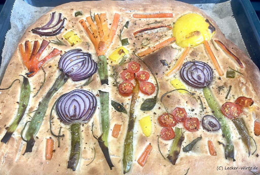 Ein Focaccia prato fiorito auf einem mit Backpapier ausgelegten Backblech, frisch aus dem Backofen. Das ligurische Fladenbrot ist mit Paprika, Frühlingszwieble, roten Zwiebelscheiben und halben Tomaten beleget und mit Salz sowie gehackten Kräutern bestreut. Es sieht aus wie eine Blumenwiese.