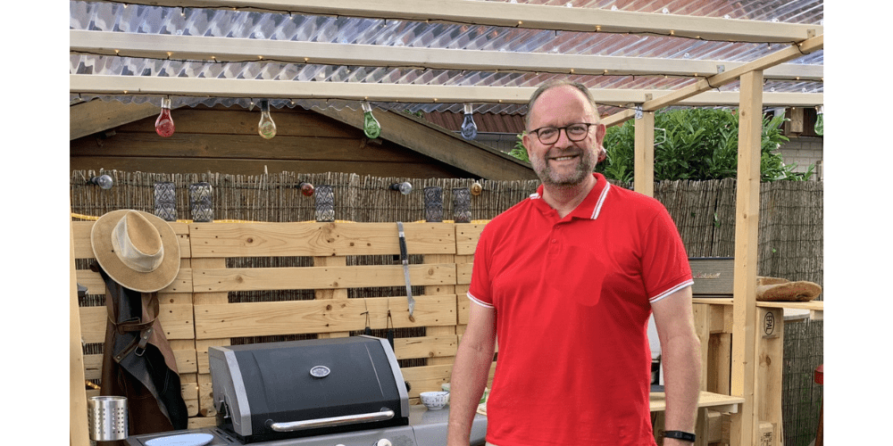 Ein Mann in knielanger, beider Hose und einem roten Poloshirt steht vor einem selbstgebauten Grillunterstand aus Paletten. Des Weiteren sieht man einen Gasgrill, ein Fass Kölsch, Barhocker, eine Gasflasche und einen Sonnenhut.