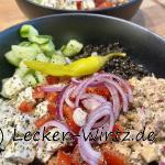 Eine Salat Bowl aus schwarzem Porzellan gefüllt mit Tomatenstückchen, mariniertem Feta, Gurkenwürfeln, Beluga Linsensalat und Thunfisch-Kichererbsen Creme, garniert mit halben Zwiebelringen und einer grünen Peperoni.