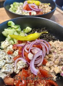 Eine Salat Bowl aus schwarzem Porzellan gefüllt mit Tomatenstückchen, mariniertem Feta, Gurkenwürfeln, Beluga Linsensalat und Thunfisch-Kichererbsen Creme, garniert mit halben Zwiebelringen und einer grünen Peperoni.