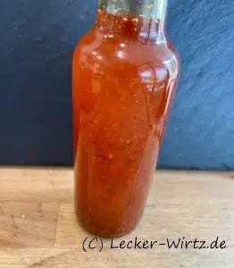Eine kleine Flasch selbstgemachtes Chiliöl ohne Etikett auf einer Holzplatte vor einem Schiefer-Hintergrund.