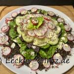 Ein sehr hübsch angerichteter Rote Bete Linsen Salat. Die Linsen liegen auf einem weißen Porzellanteller. Darüber in Schichten Salatblätter, rote Bete, Gurkenscheiben und Radieschenscheiben. Zum Schluss Dressing und geröstete Sesamkörner und als Spitze des Saltas ein kleines Salatblatt mit einer Radieschen Scheibe in der Mitte.