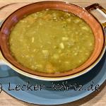 Erbsensuppe in einer roten Porzellanschüssel mit weißem Rand auf einem blauen Teller auf einem Holzbrett. Soupe aux Pois