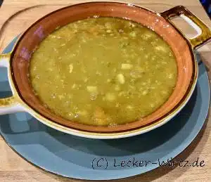 Erbsensuppe in einer roten Porzellanschüssel mit weißem Rand auf einem blauen Teller auf einem Holzbrett. Soupe aux Pois