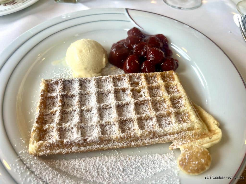 Eine Bergische Waffeln, bestreut mit Puderzucker, mit heißen Kirschen und Vanilleeis auf einem weißen Porzellanteller.