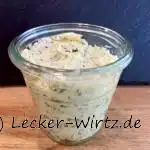 Ein kleines Weckglas mit mediterraner Parmesan-Butter auf einem Holzbrett vor einer Schiefertafel.